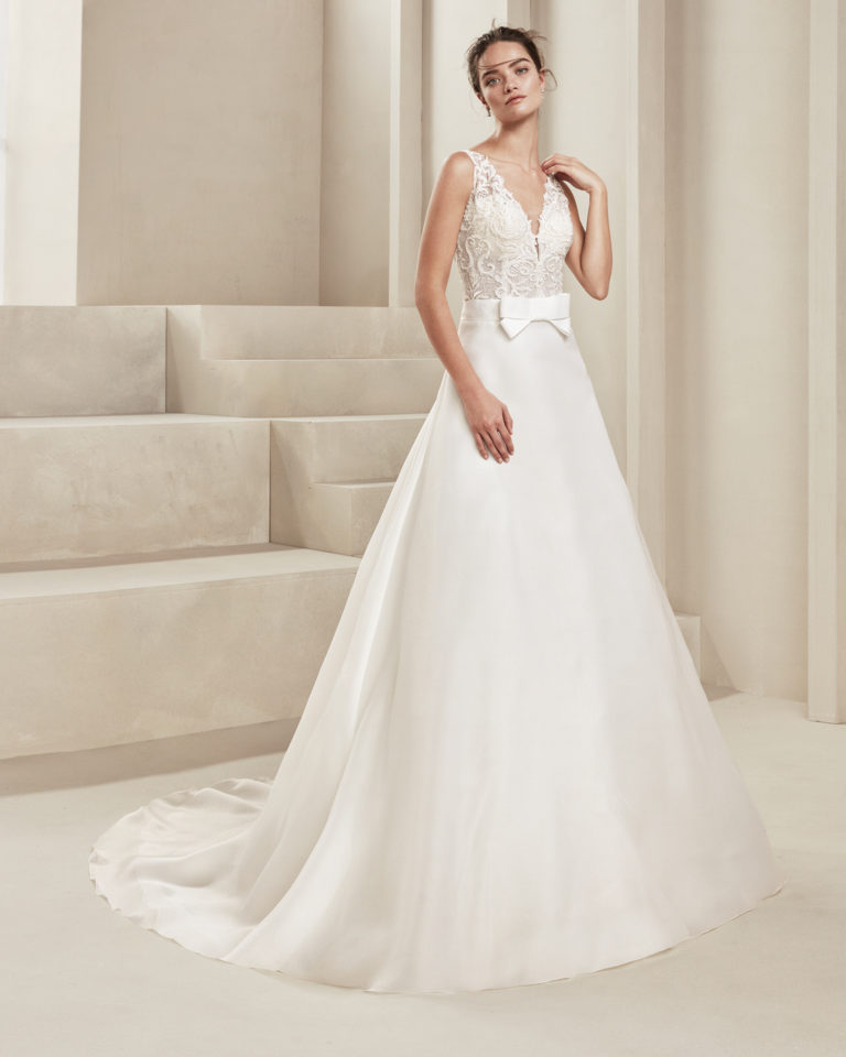 Classic wedding dress in organza and beaded lace. With deep-plunge neckline and teardrop back. With bow at the waist. Available in natural. 2019 ALMA_NOVIA Collection.