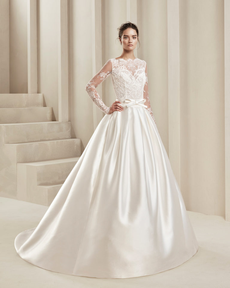 Classic wedding dress in tammy and beaded lace. With long sleeves and bow in front at the waist. Available in natural. 2019 ALMA_NOVIA Collection.