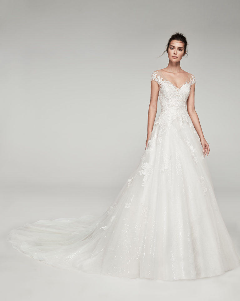 Romantic tulle and beaded lace wedding dress. With V-neckline and tulle back. Skirt with lace appliqués. Available in natural. 2019 ALMA_NOVIA Collection.