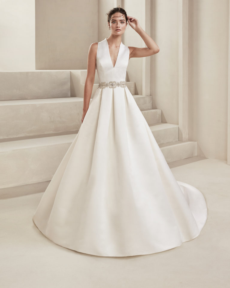 Classic ottoman wedding dress. With deep-plunge neckline and teardrop back. With beaded appliqués at the waist. Available in natural. 2019 ALMA_NOVIA Collection.