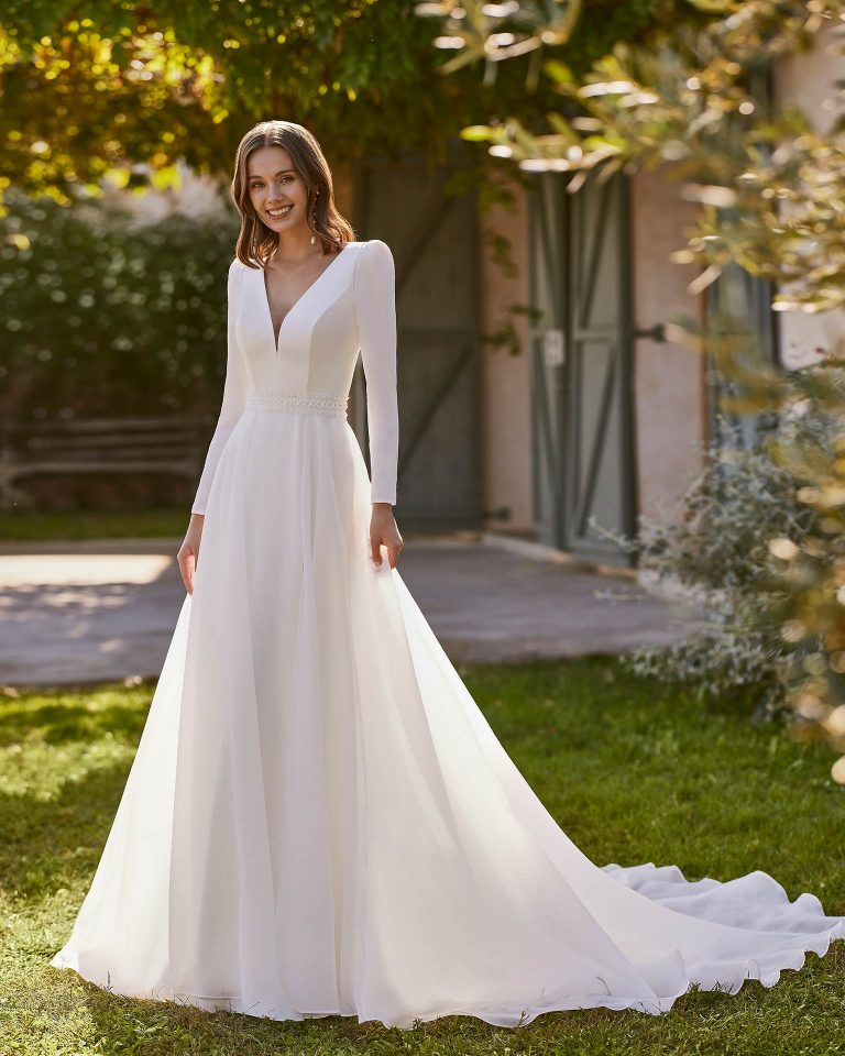 Long bridal dress, two-piece and straight cut. Crafted in crepe with a V-neckline, long sleeves, open back, and overskirt. ALMA_NOVIA.