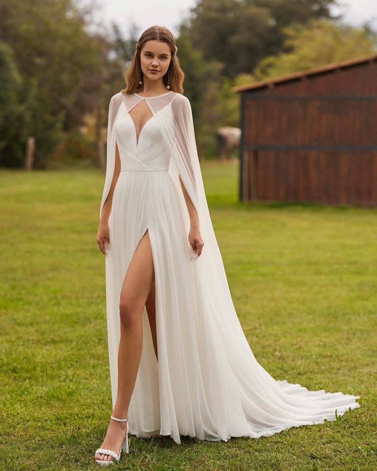 Long bridal dress, two-piece and straight cut. Crafted in silk muslin with a V-neckline, straps, open back with cape, and skirt with a side slit. ALMA_NOVIA.