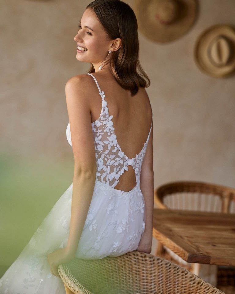 Long bridal dress with an A-line cut. Crafted in lace with a deep neckline, straps, and open back. ALMA_NOVIA.