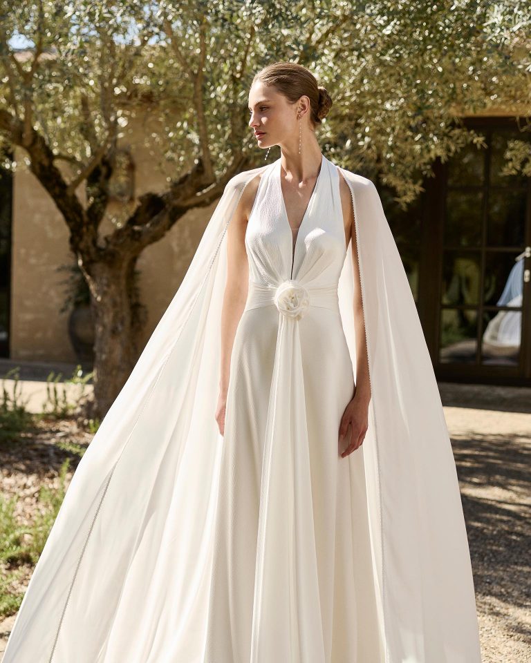 Vestido de novia de dos piezas en satén texturizado con escote halter, espalda abierta y silueta en A. Adornos florales, acabado drapeado e incluye capa de georgette. ALMA_NOVIA.